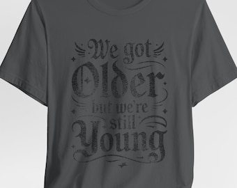 May include: Dark gray t-shirt with the text "We got Older but we're still Young" in a distressed, vintage style. The text is in a decorative font with embellishments. The shirt is made of a soft material.
