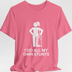 May include: Pink t-shirt with a white graphic of a person with a leg cast and crutch. The text "I DO ALL MY OWN STUNTS" is below the graphic. The shirt is made of a soft material.