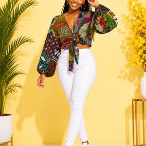 May include: A long-sleeved crop top featuring a vibrant, multi-colored patchwork pattern with a deep V-neckline and a front tie closure, paired with solid white slim-fit trousers and white athletic sneakers.