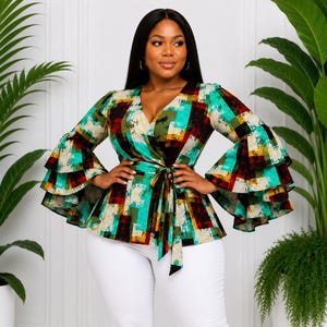 May include: A colorful wrap top with a V-neck and flared sleeves. The top features a vibrant plaid pattern in shades of green, red, yellow, and black. It is tied at the waist with a matching fabric belt. The top is paired with white pants.