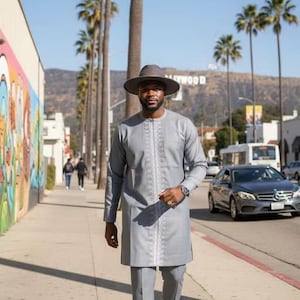 May include: A man in a light gray long-sleeve tunic and matching pants, with a gray hat and black shoes, walks on a sidewalk. The tunic has white embroidered detailing. The Hollywood sign is visible in the background.