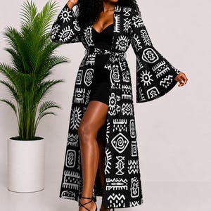 May include: A long-sleeved black duster coat featuring a white graphic print of various abstract symbols and eye motifs. The garment has a tie-waist closure, wide bell sleeves, and an open front design that reveals a solid black mini dress underneath.