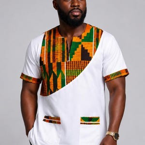 May include: A white short-sleeved shirt featuring a geometric patterned panel in orange, green, and black across the chest and shoulders, matching sleeve cuffs, two front pockets with patterned trim, and a coordinating round brimless cap.