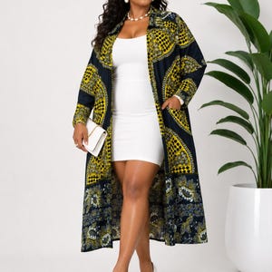 May include: A woman wearing a long, patterned coat over a white dress and white heels. The coat features a yellow and navy blue geometric design. She is holding a white clutch. A large potted plant is in the background.