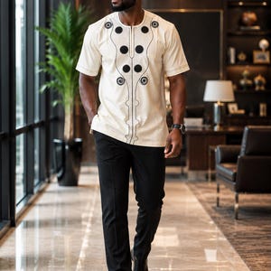 May include: A short-sleeved cream-colored shirt featuring a round neckline and black circular embroidery patterns along the chest and center front, paired with solid black trousers and black dress shoes.
