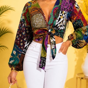 May include: A long-sleeved crop top featuring a vibrant, multi-colored patchwork pattern with a deep V-neckline and a front tie closure. The garment is paired with solid white pants, creating a high-contrast look with long, billowy sleeves.