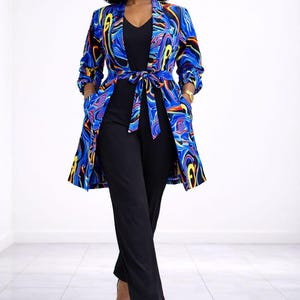 May include: A woman wearing a black jumpsuit with a long, patterned jacket. The jacket features a vibrant blue, yellow, and red abstract design. The outfit is completed with black heels.