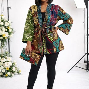 May include: A colorful patterned kimono-style jacket with a matching clutch. The jacket features a mix of floral and geometric designs in green, blue, red, and brown. The model is wearing black leggings and patterned heels.