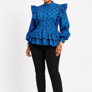 May include: A long-sleeved blue blouse featuring a black geometric pattern, a high neckline, and ruffled shoulder and waist details. The blouse is paired with slim-fit black trousers and black high-heeled pumps, creating a structured silhouette.