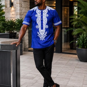 May include: A short-sleeved royal blue shirt featuring intricate white embroidery around the neckline, chest, and sleeve cuffs, paired with solid black trousers and black dress shoes. The shirt has a button-down placket and a round neckline.