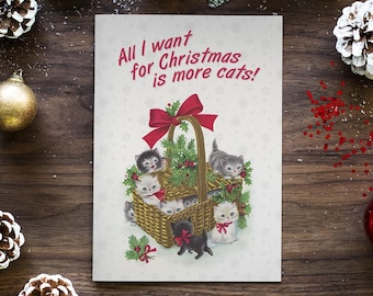Może przedstawiać: Kartka świąteczna z napisem "All I want for Christmas is more cats!" przedstawia kosz pełen kociąt. Kocięta są w różnych kolorach, a kosz ozdobiony jest czerwoną kokardą i zielenią.