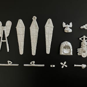 Może przedstawiać: Zestaw niepomalowanych, srebrnych metalowych części do modeli miniaturowych, w tym trzy długie, zwężające się konstrukcje przypominające skrzydła, uskrzydlona figurka, mały łukowaty otwór drzwiowy, zespół mechaniczny, śmigło oraz różne małe elementy konstrukcyjne.