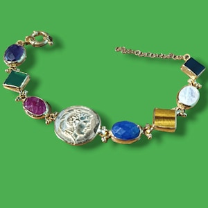 May include: A gold bracelet with a variety of gemstones, including a ruby, sapphire, emerald, amethyst, and a coin charm. The bracelet has a chain link closure.