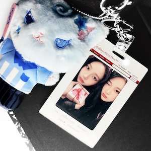 May include: A light blue plush keychain with a cartoon face, blue and pink accents, and a silver keyring. Beside it is a white ID card with two photos and the text "Archive of Our Own".