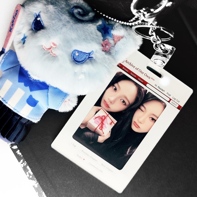 May include: A light blue plush keychain with a cartoon face, blue and pink accents, and a silver keyring. Beside it is a white ID card with two photos and the text "Archive of Our Own".