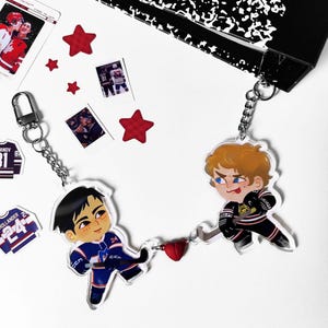 May include: A hockey-themed keychain featuring two cartoon figures in team uniforms, connected by a chain and a red heart charm. The keychain is accompanied by star-shaped stickers and small photos.