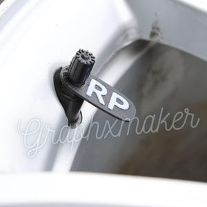 May include: Close-up of a black valve stem cap with a white "RP" label attached to a white car tyre.