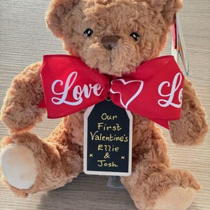 May include: A brown teddy bear with a red bow that says "Love" and a heart. The bear has a tag that says "Our First Valentine's Ellie & Josh". The bear is a plush toy.