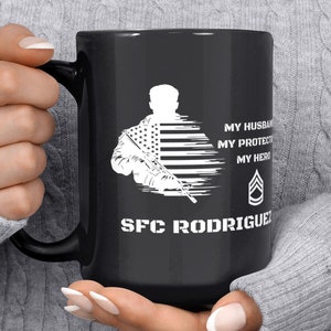 以下が含まれることがあります： 黒いセラミックマグカップ。白いグラフィックは、ライフルを持った兵士とアメリカ国旗のデザイン。テキストには「MY HUSBAND, MY PROTECTOR, MY HERO」と「SFC RODRIGUEZ」が含まれています。