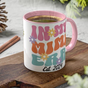 May include: White ceramic mug with a pink rim and handle. The mug features a colourful graphic that says "In My Mimi Era" with flowers and the text "Est. 2024".