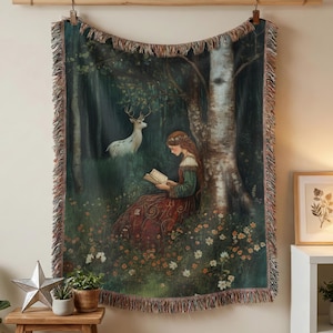A tapestry throw blanket with a woodland scene featuring a woman reading a book, a deer, and a birch tree. The tapestry has a fringed edge and is woven with a variety of colors, including green, brown, and white.
