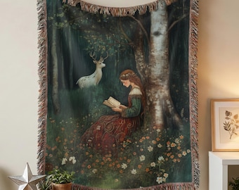 May include: A woven throw blanket featuring a printed illustration of a person in a long red dress reading a book while sitting under a large tree in a forest. A white deer stands in the background among dark green trees and small white flowers.