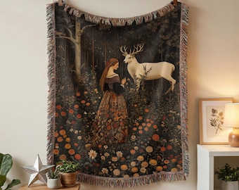 May include: A woven tapestry blanket featuring a dark forest scene with a person in a long patterned dress standing near a white deer. The blanket has a fringed edge and is displayed hanging from a wooden rod against a light wall.