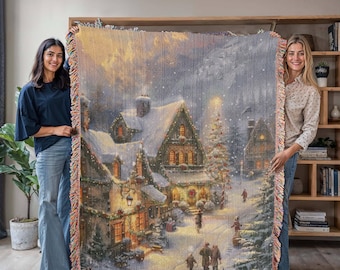 May include: A woven throw blanket featuring a winter village scene with snow-covered houses, a Christmas tree, and figures walking in the snow. The blanket has a fringed edge and is held up by two people.