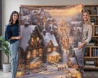 May include: A woven tapestry blanket featuring a winter village scene with snow-covered buildings, a decorated Christmas tree, and warm lighting. The blanket has a fringed edge and is held up by two people.