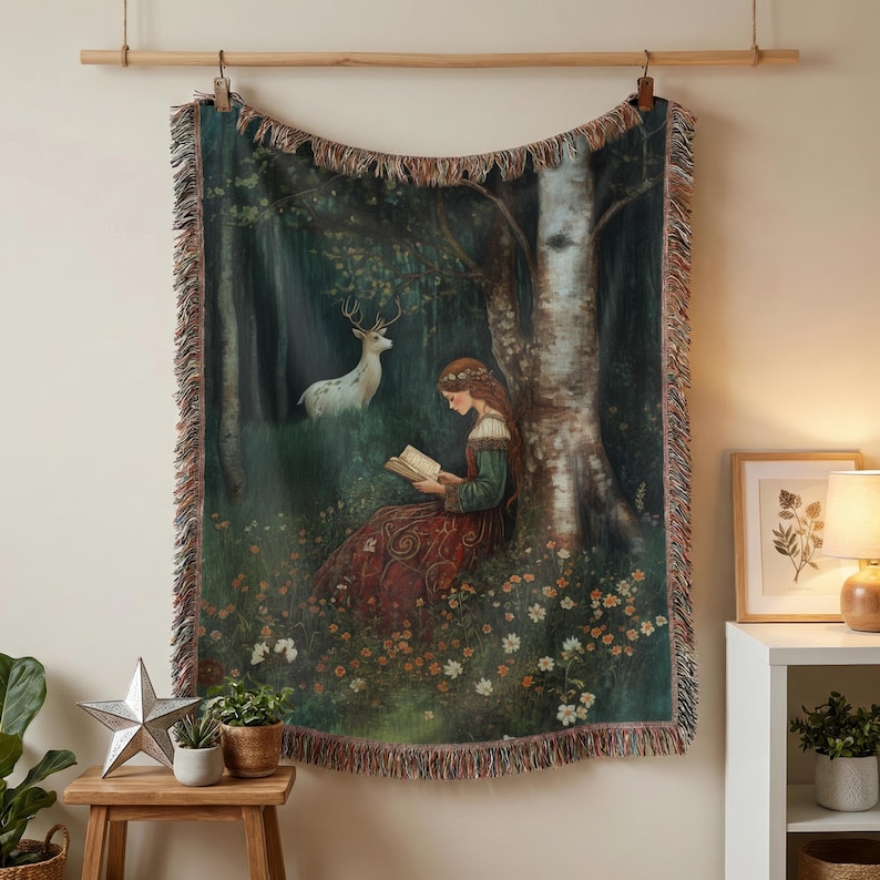 A tapestry throw blanket with a woodland scene featuring a woman reading a book, a deer, and a birch tree. The tapestry has a fringed edge and is woven with a variety of colors, including green, brown, and white.