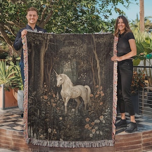 K&ouml;nnte beinhalten: Eine Wandteppich-Decke mit einem wei&szlig;en Einhorn, das in einem dunklen Wald mit floralem Muster steht. Der Wandteppich ist mit einem Fransenrand gewebt.