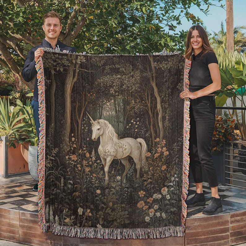 K&ouml;nnte beinhalten: Eine Wandteppich-Decke mit einem wei&szlig;en Einhorn, das in einem dunklen Wald mit floralem Muster steht. Der Wandteppich ist mit einem Fransenrand gewebt.