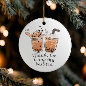 May include: A round white ceramic ornament featuring a graphic illustration of two bubble tea cups with brown liquid and black tapioca pearls. The text "Thanks for being my best-tea" is printed in black below the cups. It hangs from a gold string.