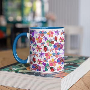 May include: White ceramic mug with a blue handle and interior. The mug features a vibrant floral pattern in pink, purple, and orange. It rests on a book with a floral design. The mug is suitable for tea or coffee.