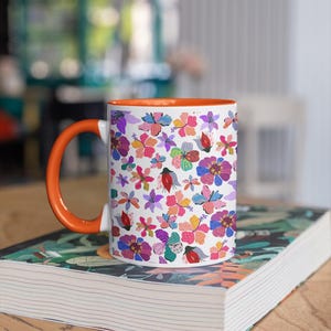 May include: White ceramic mug with a vibrant floral pattern in pink, purple, and orange. The mug has an orange handle and interior, resting on a book with a floral cover. A colorful and cheerful design for a coffee or tea cup.