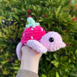May include: A pink crocheted turtle with a pink and white crocheted strawberry shell. The turtle has black eyes and a green crocheted leaf on top of the strawberry.