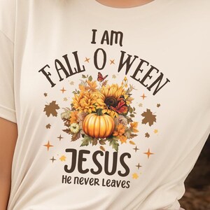 Pode incluir: Camiseta creme com o texto "I AM FALL O WEEN JESUS He never leaves". A camiseta apresenta um gráfico com tema de outono com uma abóbora, girassóis e folhas de outono. Abóboras estão no fundo.