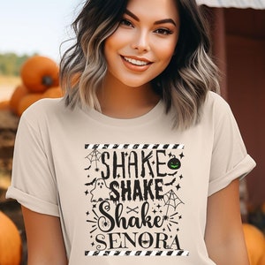 May include: A beige t-shirt with a black graphic print that says "Shake Shake Shake Senora" with Halloween-themed imagery like ghosts, bats, spiders, and a pumpkin.