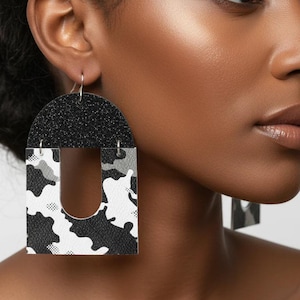 May include: Black and white camouflage print earrings with a glittery black top section. The earrings are a unique U-shape design, with a silver hook. The earrings are a bold fashion statement.