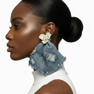 May include: A statement earring featuring a white floral-shaped stud with a cluster of small pearls at the center, attached to a large, triangular piece of distressed blue denim fabric with frayed edges that hangs down from the earlobe.
