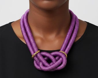 May include: A vibrant purple necklace with a knotted design. The necklace is made of thick, textured material and features two gold-colored rings. The necklace is worn against a black top.
