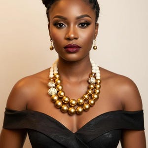 Könnte beinhalten: Eine Frau in einem schwarzen schulterfreien Kleid und Goldschmuck. Die Halskette besteht aus großen goldenen und weißen Perlen. Die Ohrringe sind kleine goldene Kugeln. Ihr Haar ist zu einem Dutt frisiert.