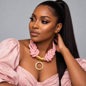 May include: A statement necklace featuring thick, soft pink knotted rope segments connected by a series of polished gold-toned cylindrical beads, centered with a large, circular gold-toned pendant with a textured, hammered finish.