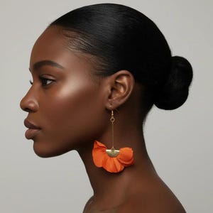 Puede incluir: Perfil lateral de una persona que lleva pendientes de flores naranjas. Los pendientes tienen una base de semicírculo dorada y un largo alambre dorado. Los pétalos de la flor son de color naranja vibrante.