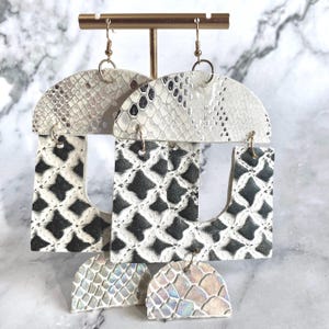 Pode incluir: Tr&ecirc;s pares de brincos pendurados com um design geom&eacute;trico. As se&ccedil;&otilde;es superior e inferior apresentam um padr&atilde;o de pele de cobra em tons de branco e cinza. A se&ccedil;&atilde;o do meio apresenta um padr&atilde;o geom&eacute;trico em preto e branco. Os brincos s&atilde;o suspensos por ganchos dourados.