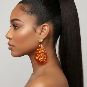 May include: Gold-toned earrings with a textured circular stud and a large, teardrop-shaped pendant. The pendant is filled with sparkling gold glitter, creating a glamorous and eye-catching accessory.