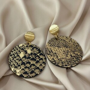 Op de afbeelding: Een paar goudkleurige oorbellen met een cirkelvormig ontwerp. De oorbellen hebben een textuur, slangenleerpatroon in zwart en goud. De oorbellen hebben een kleine, ronde knop aan de bovenkant.