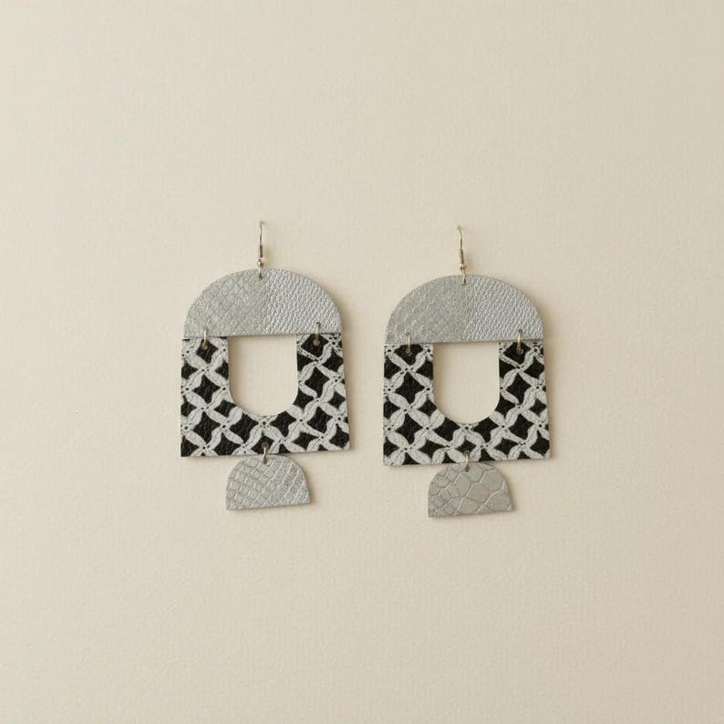 Pode incluir: Um par de brincos geom&eacute;tricos prateados e pretos. Os brincos apresentam uma parte superior prateada em semic&iacute;rculo, um centro com padr&atilde;o a preto e branco e uma pequena parte inferior prateada em semic&iacute;rculo. Os brincos est&atilde;o pendurados em ganchos prateados.