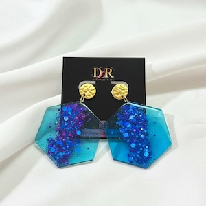 May include: Hexagonal earrings in teal and blue, with gold studs. The earrings are filled with blue and purple glitter. They are displayed on a black card with the text "DR" and "www.Designdby.com".