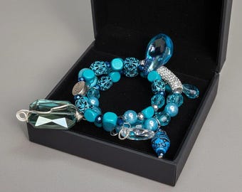 May include: A turquoise beaded bracelet with various shapes and sizes of beads, including faceted crystals and decorative charms, rests inside a black jewelry box. The bracelet features a mix of light and dark blue tones, with silver accents.